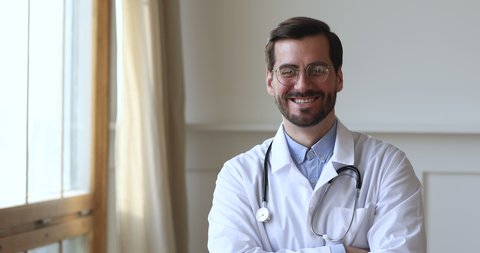 Smiling young man professional medic doctor wear white medical uniform glasses looking at camera standing in hospital office, happy friendly male gp physician surgeon doc close up view portrait