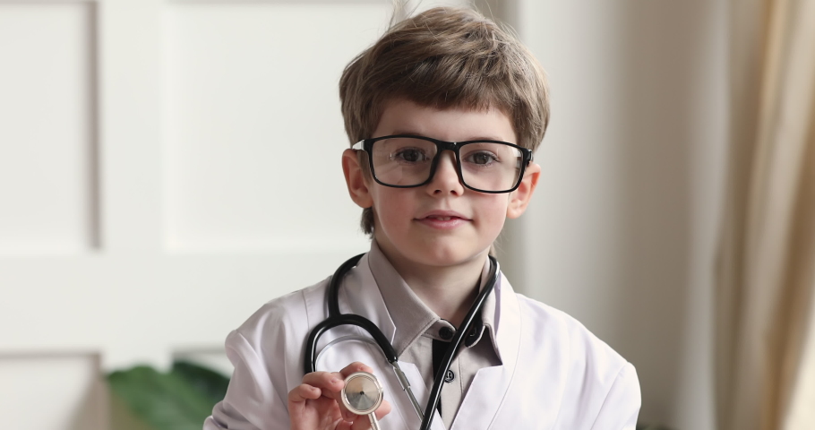 Happy funny adorable cute little preschool child boy wear white medical coat glasses holding stethoscope looking at camera playing doctor alone, children future professions concept, closeup portrait Royalty-Free Stock Footage #1044921055