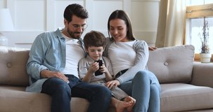 Smiling parents and child son using smart phone app enjoy modern gadget tech relax together sit on sofa, happy family of three look at cellphone doing shopping in mobile application on couch at home