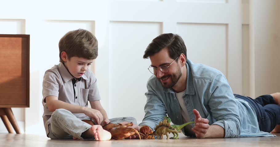 Loving young adult dad and cute preschool child son talking playing dinosaurs toys sit on warm floor, happy single parent father helping little kid explain paleontology having fun at home together Royalty-Free Stock Footage #1044921073