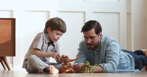 Loving young adult dad and cute preschool child son talking playing dinosaurs toys sit on warm floor, happy single parent father helping little kid explain paleontology having fun at home together