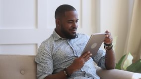 Smiling biracial young guy enjoying leisure weekend time, using digital tablet at home. Happy african american man playing online games, watching funny videos, swiping photos, chatting with friends. - Powered by Shutterstock - Get 15% off with code: PIKWIZARD15