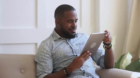 Smiling biracial young guy enjoying leisure weekend time, using digital tablet at home. Happy african american man playing online games, watching funny videos, swiping photos, chatting with friends.