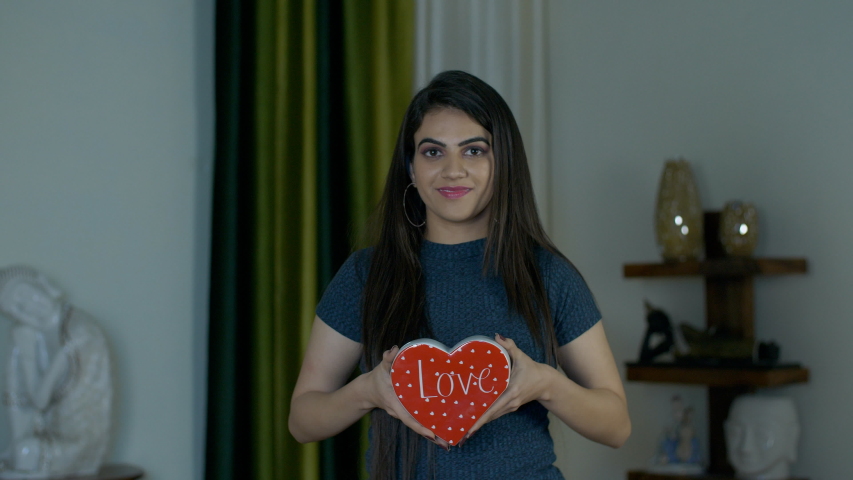 Pretty Indian girl smiling and showing a red heart symbol of love to the camera. Young happy female holding a ceramic heart-shaped toy with a wide toothy smile on her face on Valentine