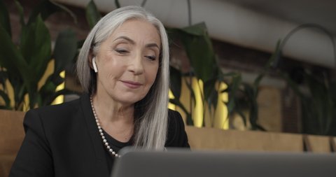 Close up of old businesswoman in earpods holding having videocall on her laptop and smiling. Mature modern woman sitting in cafe and communicating and waving to computer camera.