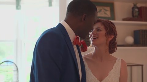 A bride and groom in the living room stand close and smile at each other