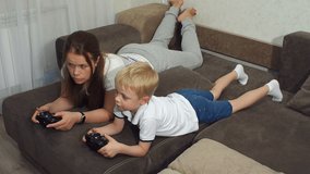 A young mother with a small blond son playing computer games lying on the couch at home, they are holding joysticks. Slow motion. Woman with child playing computer game.
