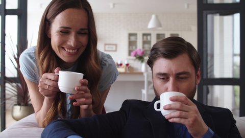 Closeup smiling couple drinking tea at home together. Happy woman bringing cups of tea for husband in living room. Portrait of joyful couple taking coffee break on sofa in slow motion.