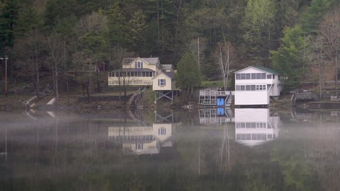 Mist gently drifts across the top of a glassy still lake reflecting the surrounding houses and forrest covering the hills on a relaxing calm day.
