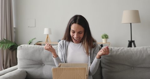 Amazed young woman opening cardboard box with internet store order, excited by successful purchase. Happy mixed race girl receive carton package with unexpected gift, sitting in living room at home.