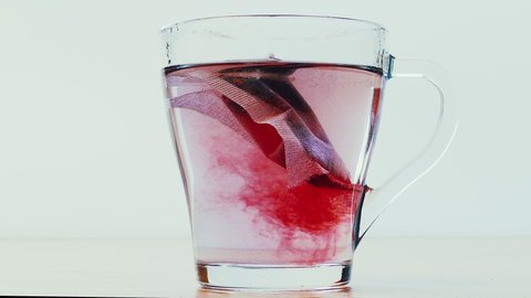 In a glass cup with a bag of fruit tea pour boiling water. Glass mug of tea made from the petals of hibiscus isolated on light background. Glass cup of herbal tea close-up