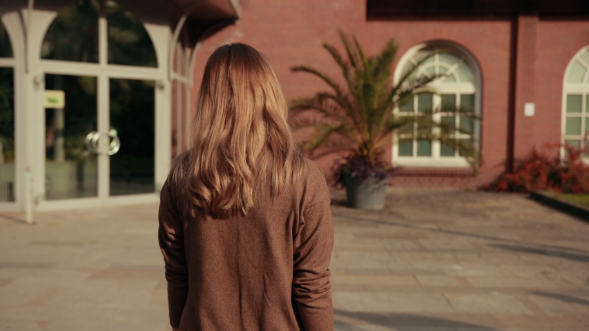 Young woman with brown hair brown sweater walks away from camera, twists around, smiles calls gestures with her index finger. Behind beautiful Park with palm trees trees house