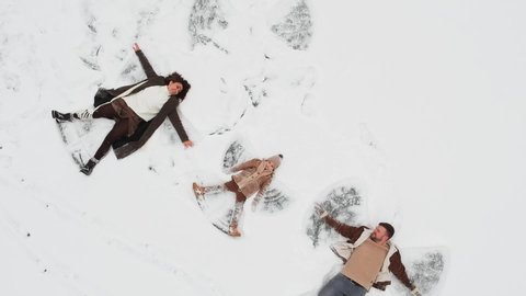 Aerial view. Happy Family couple with young child kid of three people lies on snow and makes snow angel and the camera moves away up and around, frozen lake in USA, Alaska. Chocolate brown clothing.
