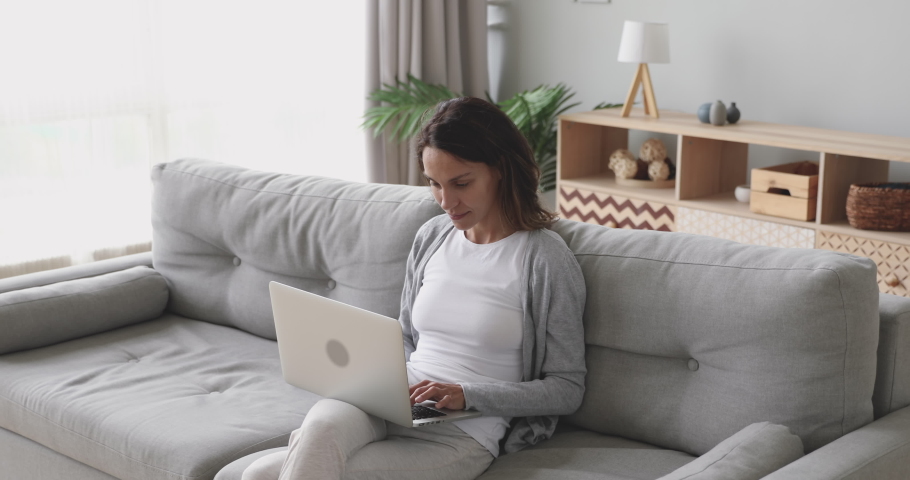 Focused smiling young mixed race woman sitting on sofa with computer, working online. Happy female freelancer enjoying distant job at home. Concentrated student studying or making purchases. Royalty-Free Stock Footage #1044943243