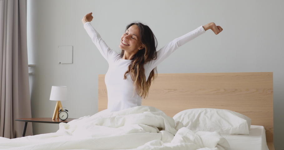 Happy mixed race girl waking up in morning after good night sleep, feeling energetic, stretching arms and back. Smiling millennial woman sitting on bed, feeling refreshed, full of energy in bedroom. Royalty-Free Stock Footage #1044943255