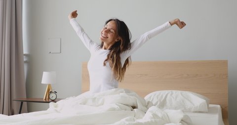 Happy mixed race girl waking up in morning after good night sleep, feeling energetic, stretching arms and back. Smiling millennial woman sitting on bed, feeling refreshed, full of energy in bedroom.
