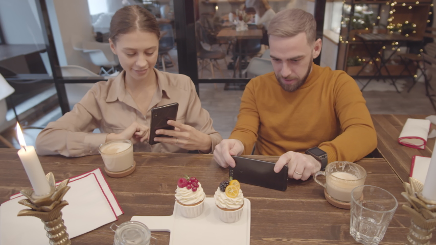 Medium shot of young Caucasian man and woman sitting at café table and making photos of cupcakes using smartphones camera