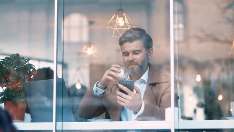 Distant view of handsome middle aged businessman with grey hair works drinks coffee smiles in cafe. Business and entrepreneurship. Successful, confident, social network, apps, laptop.