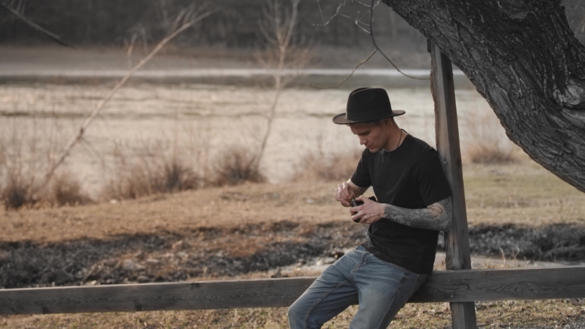 A young attractive brutal male photographer in a hat with a tattoo on his arm adjusts the camera sitting in the open air near a tree. Warm autumn shades. Slow motion, 4K, Red Epic