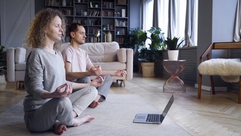 Happy couple in pajamas doing morning practice in modern living room. Side view woman and man sitting in lotus pose on carpet meditates before laptop while watching spiritual practices lessons 