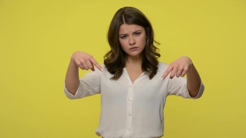 Bossy attractive brunette woman in blouse pointing down with serious demanding expression, gesturing here and right now, showing direction below. indoor studio shot isolated on yellow background