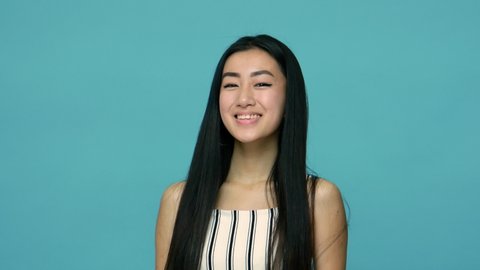 Charming positive asian woman with long straight black hair winking playfully and smiling flirting at camera, having some cunning idea, blinking eye. indoor studio shot isolated on blue background