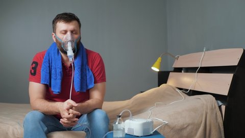 Man makes himself inhalation using nebulizer. Young man sits alone in room with mask for inhalation on his face and breathes sprayed saline. Slow motion and general plan