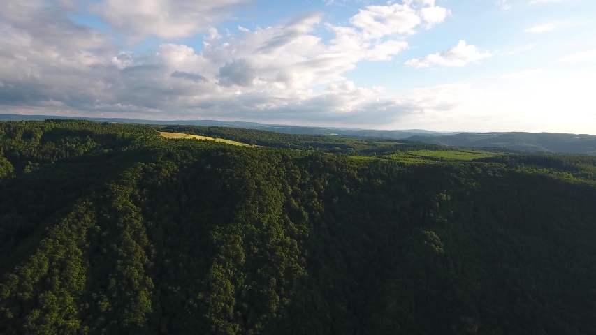 High forward flying drone aerial view of a forest overgrown hill and fields bellow