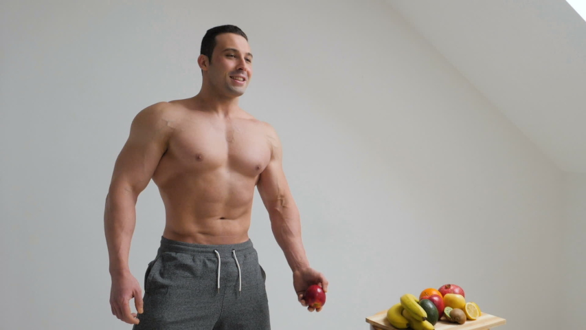 Handsome bodybuilder throws fresh fruits and posing on white background. Organic food and health concept