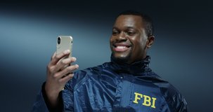 African American young happy and cheerful handsome man in FBI uniform smiling and having videochat on the smartphone via web cam on the dark wall background.