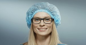 Close up of the Caucasian pretty female young doctor in glasses, blue hat smiling cheerfully to the camera. Portrait shot.