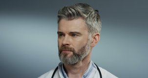 Portrait of the Caucasian good looking serious grey-haired doctor looking at the side and turning face to the camera on the gray wall background. Close up.