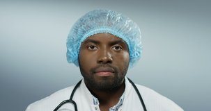 Close up of the young African American handsome man doctor looking straight to the camera with a serious face on the grey wall background. Portrait.
