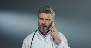 Caucasian good looking grey-haired male medic in white gown speaking on the mobile phone on the gray background. Close up.