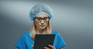 Young blonde Caucasian pretty woman doctor in blue hat and glasses using tablet device, tapping and scrolling on it.