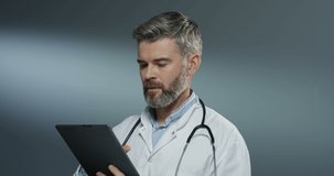 Caucasian handsome male physician in white gown using tablet device, tapping and scrolling on the gray wall background.