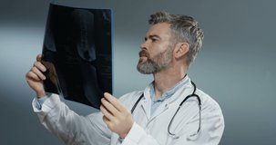Caucasian gray-haired good looking man doctor holding X-ray copy and studying it carefully on the grey background.