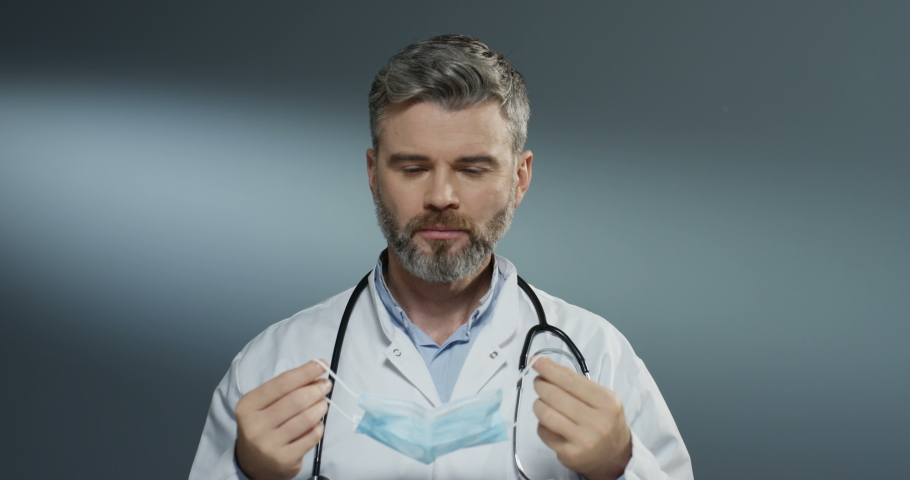 Caucasian good looking man surgeon taking on blue mask when getting ready for the surgery on gray wall background. Close up.