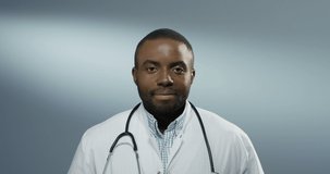 Close up of the African American young happy male doctor or intern smiling cheerfully to the camera on the gray wall background. Portrait.