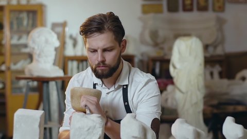 Attractive concentrated 30-aged creative bearded sculptor knocking off from chalk-stone needed shape of future sculpture using hammer and chisel