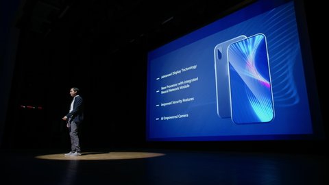 Live Event with Brand New Products Reveal: Keynote Speaker Presents Smartphone Device to the Audience. Movie Theater Screen Shows Mock-up Touch Screen Mobile Phone with High-Tech Features, Highlights