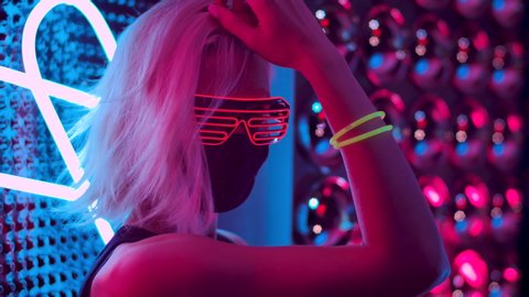Portrait One 30s Expressive Woman at Party In Place With Purple Blue Lighting. Positive Woman Closeup Face in Mask and Glow Eyeglasses Standing Confident in Colourful Neon Light and Looking at Camera