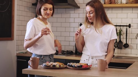 Two funny young lesbians eat sushi rolls at home in the kitchen. A woman carefully feeds her partner.