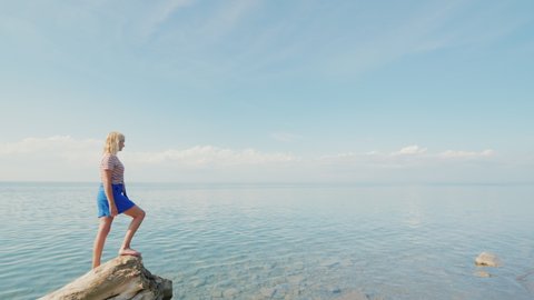 Active woman traveler stands on the edge of a log, admires a beautiful view of the calm sea and blue sky