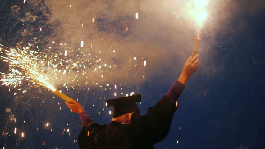 An emotional college graduate in a mantle and a graduation cap with two fireworks in his hands Royalty-Free Stock Footage #1045039942