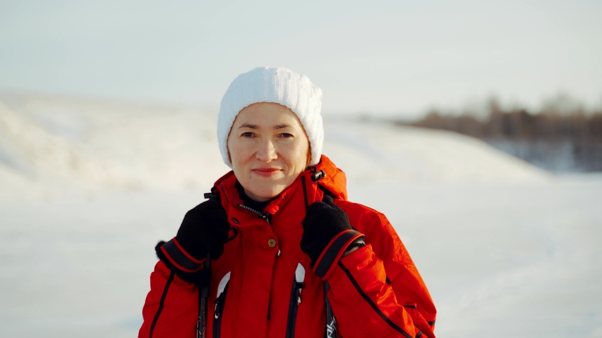 A woman in a red jacket and white hat looks into the camera. Sunny winter day.Concept of: winter holidays, family, christmas, skiing.