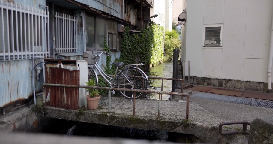 Rural Japanese housing structure with waterway channel flowing under parked bike behind residential housing property.