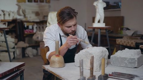 Slow motion of 30-aged creative bearded stonemason which sitting in the workshop and scraping the remains of chalk-stone using brush