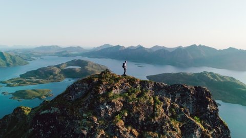 4K slow motion drone shot with orbit motion of young Norwegian climber with backpack standing on top of tall cliff admiring the panoramic sunset view in Lofoten Vesterålen in Norway.