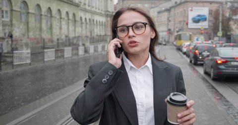 Busy young female Office Worker running in the Street while speaking on Smartphone. Holding paper cup with Coffee, hurrying up to her Office Building. Businesswoman is late for her Business Meeting